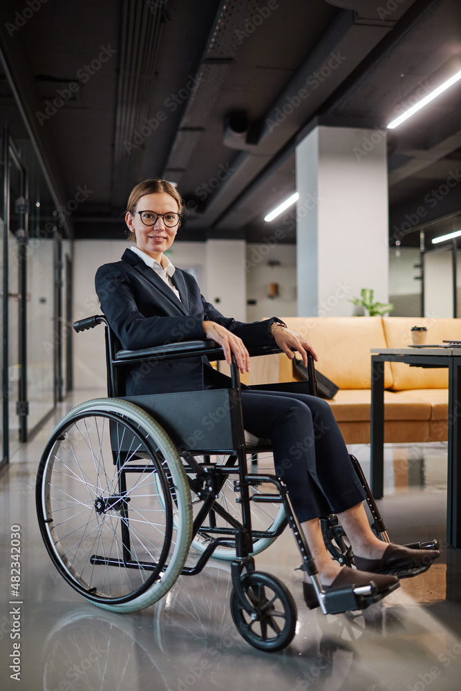 Vertical full length portrait of successful businesswoman using wheelchair and looking at camera smiling while posing in modern office space Vertical full length portrait of successful businesswoman using wheelchair and looking at camera smiling while posing in modern office space