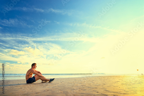 Relaxation after sports. Young muscular man sitting on the sea beach enjoying sunset.