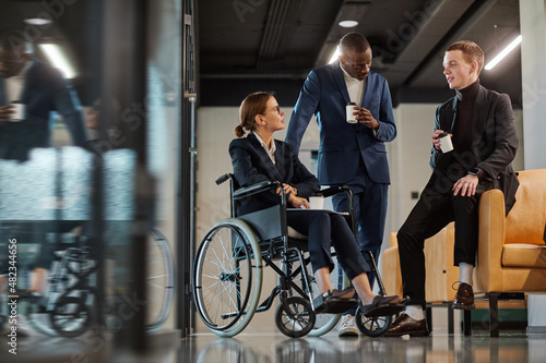 Full length portrait of diverse business team with young woman in wheelchair chatting to male colleagues in modern office