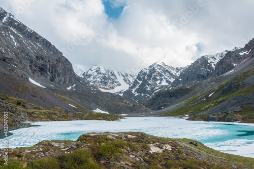 Atmospheric highland landscape with frozen alpine lake and high snowy mountains. Awesome scenery with icy mountain lake on background of large snow mountains in low clouds. Scenic view to ice lake.