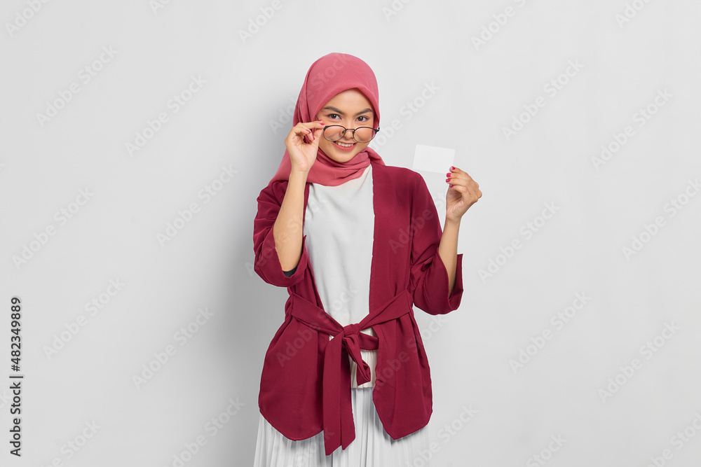 Smiling beautiful Asian woman in a casual shirt and hijab showing credit card, taking off glasses isolated over white background. People religious lifestyle concept