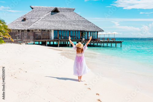 middle-aged woman plus size model in white long beach cape and sun hat walks along picturesque sandy beach in Maldives island near water villa, concept of luxury travel