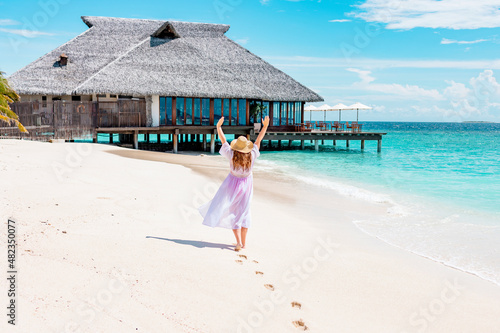 middle-aged woman plus size model in white long beach cape and sun hat walks along picturesque sandy beach in Maldives island near water villa, concept of luxury travel