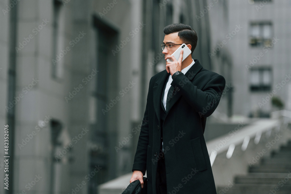 Talking by phone. Businessman in black suit and tie is outdoors in the city