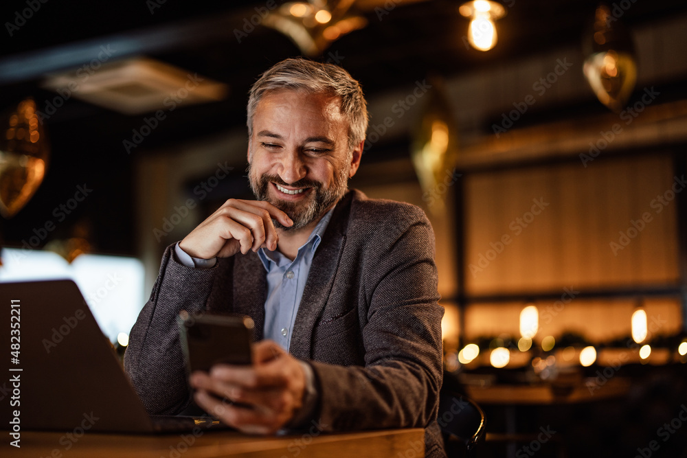 Smiling caucasian man, comparing notes on his laptop, with notes on his phone.