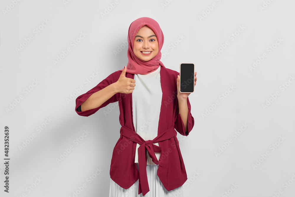 Smiling beautiful Asian woman in casual shirt and hijab holding a mobile phone with a blank screen, showing thumb up isolated over white background. People religious lifestyle concept