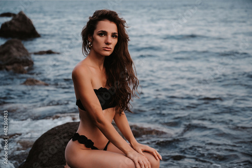 young girl in a black swimsuit is sitting sexy on the rocks on the beach by the sea in summer on vacation