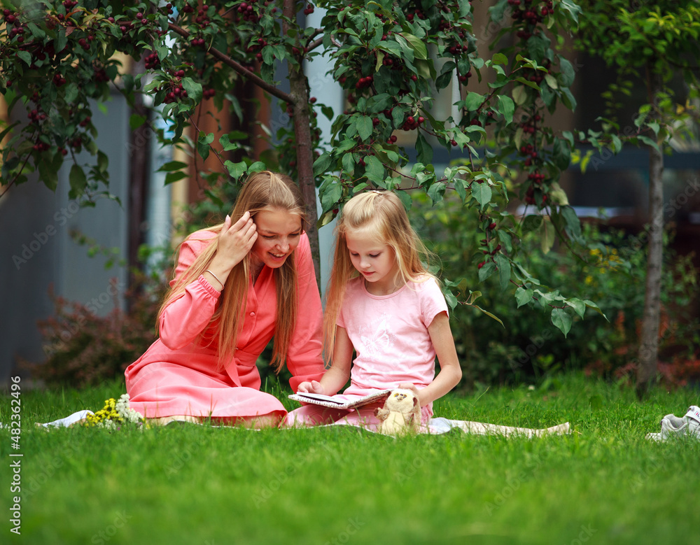 Naklejka premium Mother and daughter on green grass lawn reading book together in backyard garden