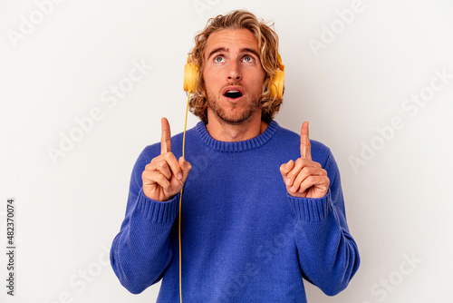 Young caucasian man listening to music isolated on white background pointing upside with opened mouth.
