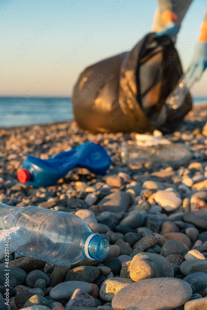 Fototapeta premium Close-up of plastic bottles against the background of a young woman with a black garbage bag on a pebble beach of the sea at sunset.Concept of ecology,volunteering.Vertical photo