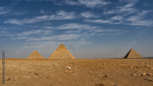 The three great pyramids of Giza on a background of blue sky and clouds. The stones are scattered on the desert sand. Silhouettes of skyscrapers of modern Cairo are visible on the horizon  in the haze