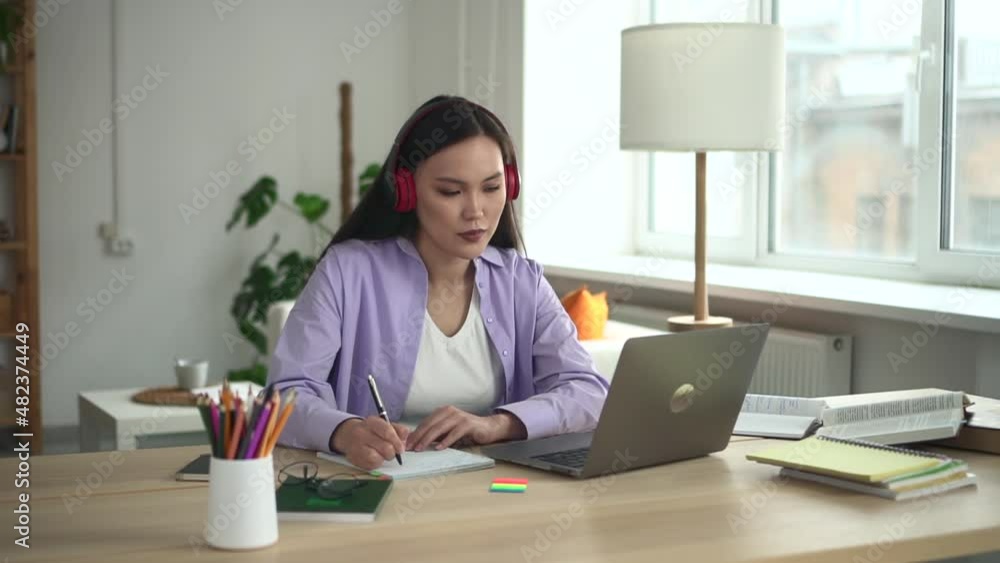 Beautiful woman student studying online and sitting at table with laptop in home room spbd. Asian young female looks at computer screen and writes in notebook, listens to lecture with headphones and Beautiful woman student studying online and sitting at table with laptop in home room spbd. Asian young female looks at computer screen and writes in notebook, listens to lecture with headphones and