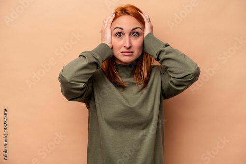Young caucasian woman isolated on beige background covering ears with hands trying not to hear too loud sound. © Asier