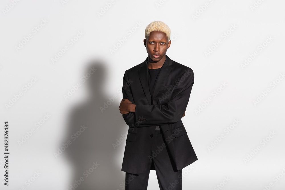blonde african american man in black suit standing with crossed arms on grey with shadow. blonde african american man in black suit standing with crossed arms on grey with shadow.