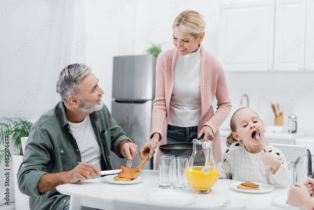 cheerful woman serving pancakes for husband during breakfast with grandchildren.