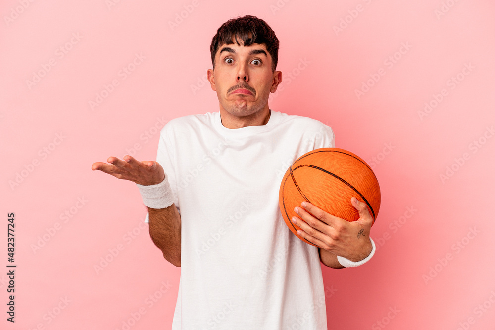 Young mixed race man playing basketball isolated on pink background shrugs shoulders and open eyes confused.