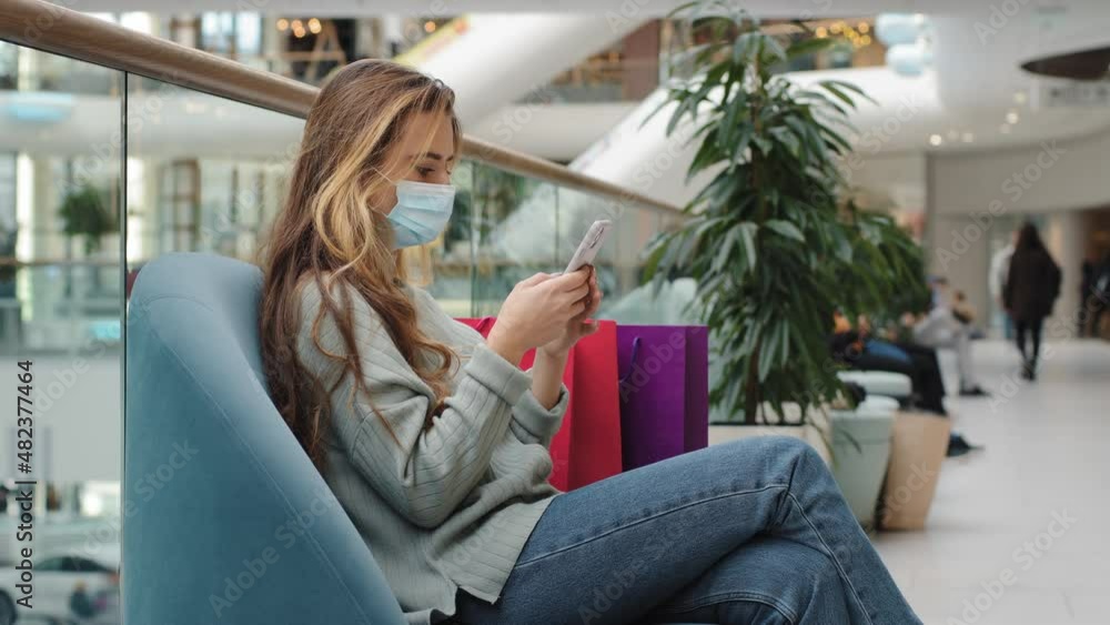 Caucasian woman buyer wearing medical mask relaxed with shopping bags female ill shopper sitting indoors on sofa in store looking at mobile phone buying online app choosing product chatting free wifi Caucasian woman buyer wearing medical mask relaxed with shopping bags female ill shopper sitting indoors on sofa in store looking at mobile phone buying online app choosing product chatting free wifi