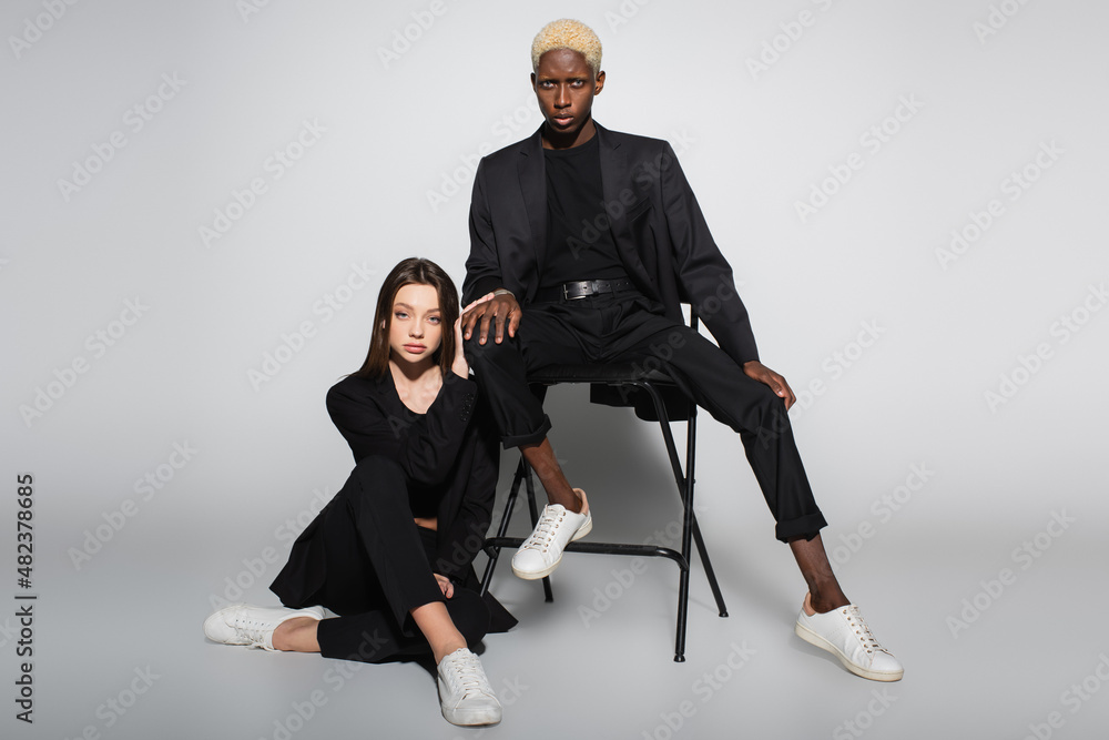 blonde african american man sitting on chair near stylish woman on floor on grey with shadow. blonde african american man sitting on chair near stylish woman on floor on grey with shadow.