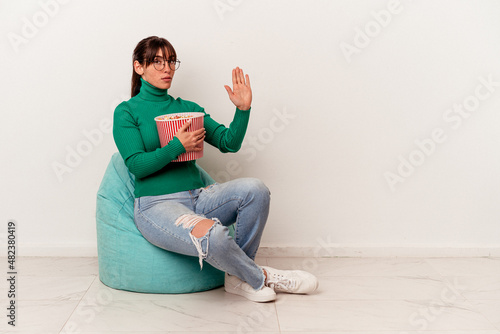 Young Argentinian woman eating popcorns on a puff isolated on white background standing with outstretched hand showing stop sign, preventing you. photo