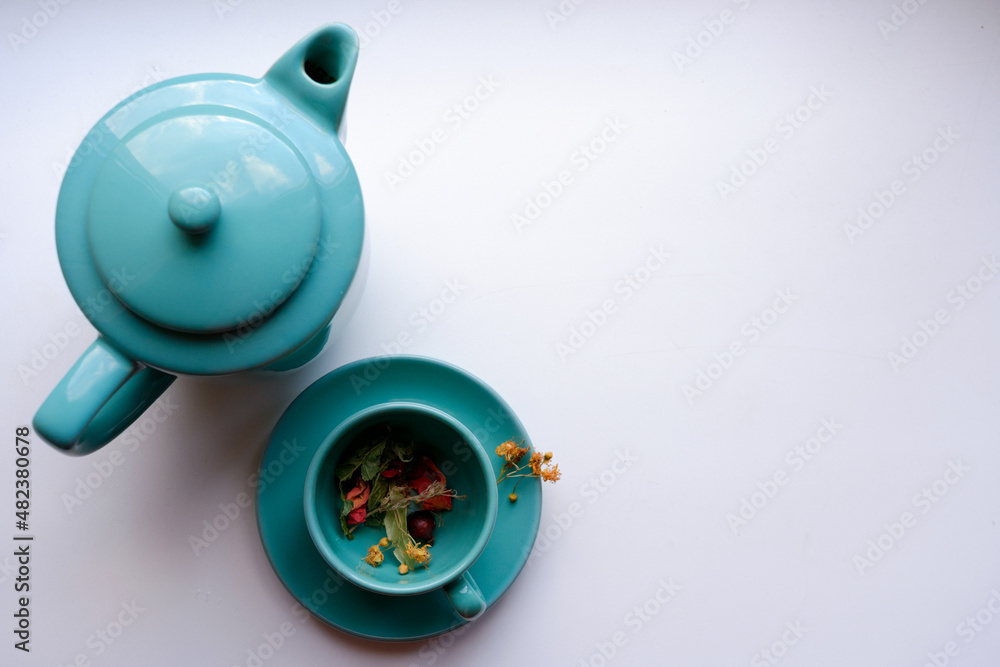 Top view: a mug on a saucer with colored herbs for tea and a teapot on a white background Top view: a mug on a saucer with colored herbs for tea and a teapot on a white background