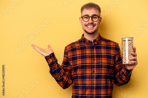 Young caucasian man holding chickpeas isolated on yellow background showing a copy space on a palm and holding another hand on waist.