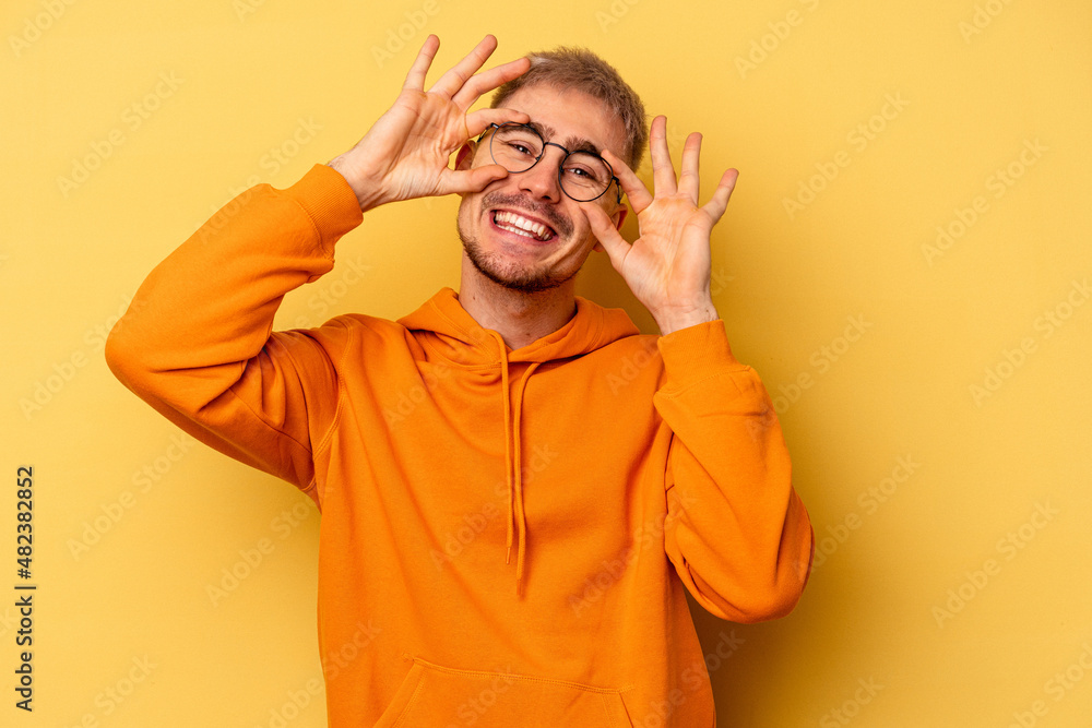 Young caucasian man isolated on yellow background showing okay sign over eyes Young caucasian man isolated on yellow background showing okay sign over eyes