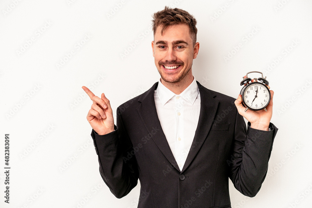 Young caucasian business man holding alarm clock isolated on white background smiling and pointing aside, showing something at blank space. Young caucasian business man holding alarm clock isolated on white background smiling and pointing aside, showing something at blank space.