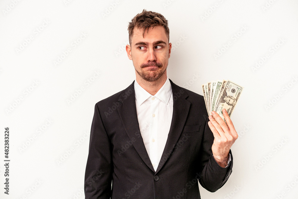 Young caucasian business man holding bank notes isolated on white background confused, feels doubtful and unsure. Young caucasian business man holding bank notes isolated on white background confused, feels doubtful and unsure.