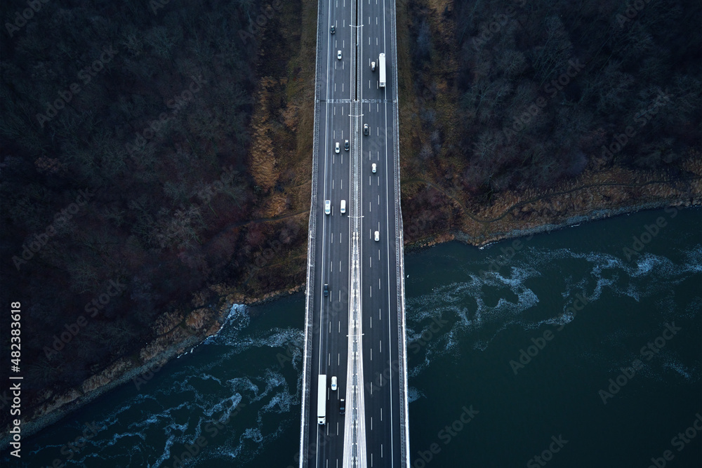 Car traffic moving on highway bridge, aerial view. Countryside road through autumn forest over river. Road trip, delivery and logistic concept
