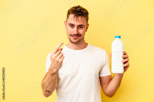 Young caucasian man holding a bottle of mil isolated on yellow background pointing with finger at you as if inviting come closer.