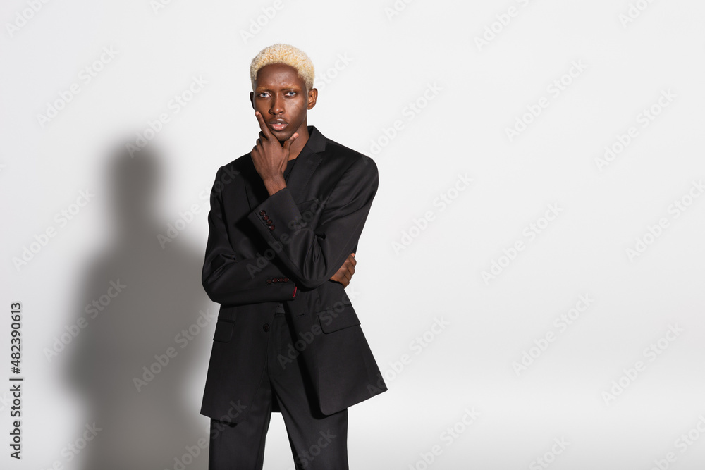 blonde african american man in black suit holding hand near face while posing on grey with shadow. blonde african american man in black suit holding hand near face while posing on grey with shadow.