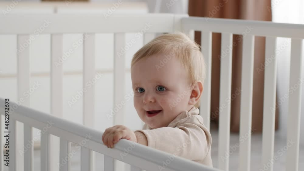 Cute Infant Kid With Rattle Toy In Hand Standing In Baby Bed Cute Infant Kid With Rattle Toy In Hand Standing In Baby Bed