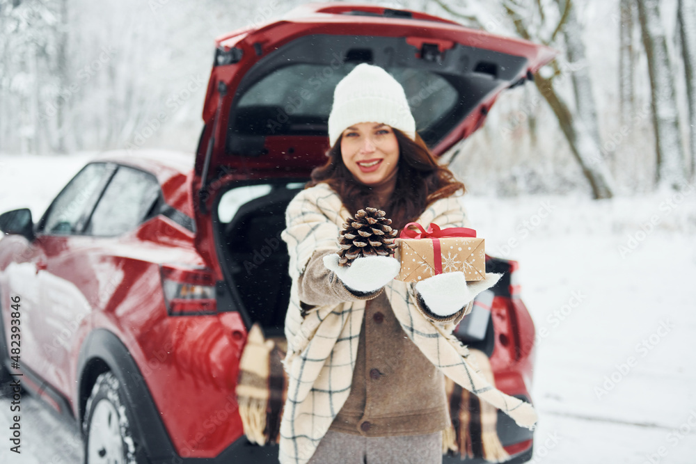 Holding gift box. Beautiful young woman is outdoors near her red automobile at winter time
