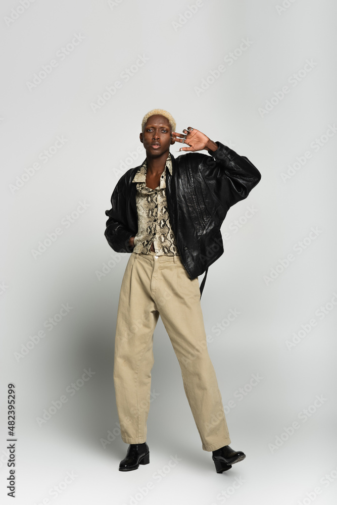 full length view of blonde african american man in black jacket and beige trousers looking at camera on grey. full length view of blonde african american man in black jacket and beige trousers looking at camera on grey.
