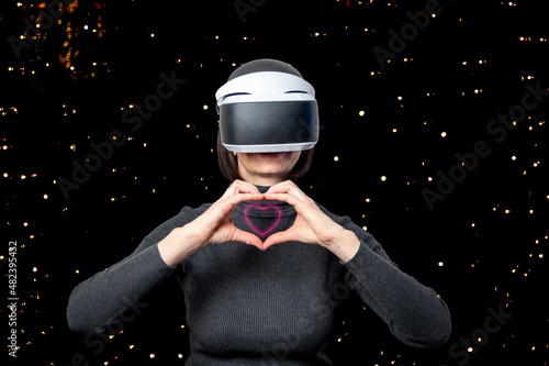 Beautiful woman using vr headset shows glowing heart shape with hands on dark background with bokeh. The concept of virtual relationships. Virtual love concept.