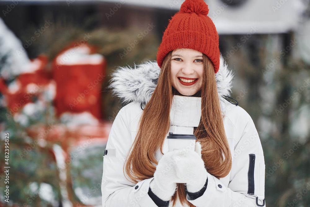 Obraz premium With cup of drink in hands. Happy young woman standing outdoors and celebrating christmas holidays