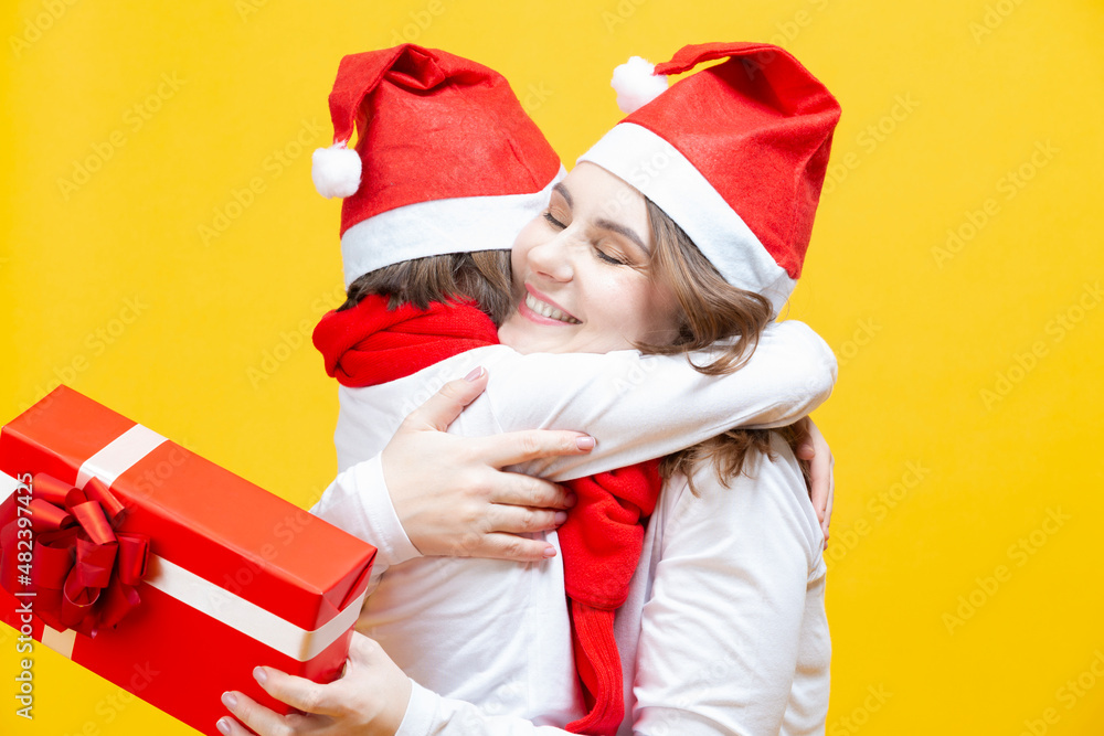 Fototapeta premium Portrait of Two Beautiful Caucasian Ladies of Different Ages Embracing Together in Santa Hats With Wrapped Red Giftbox Behind and Casual Clothes Against Trendy Yellow Background. Vertical Image