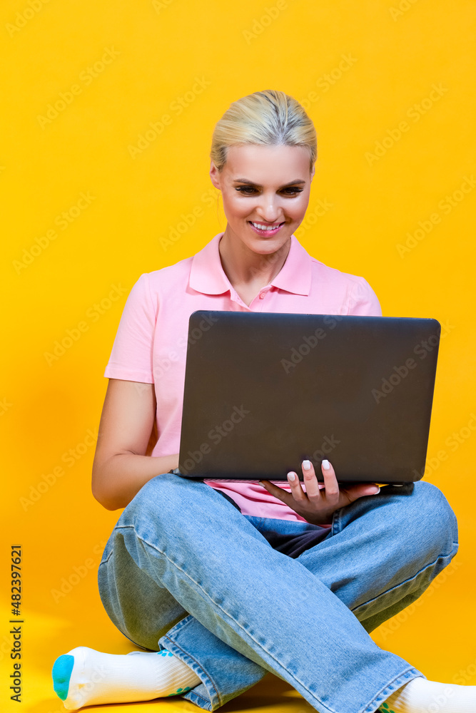 Naklejka premium Portrait of Smiling Caucasian Blond Girl with Laptop Posing With Smile Sitting on Floor Against Colorful Yellow Background.