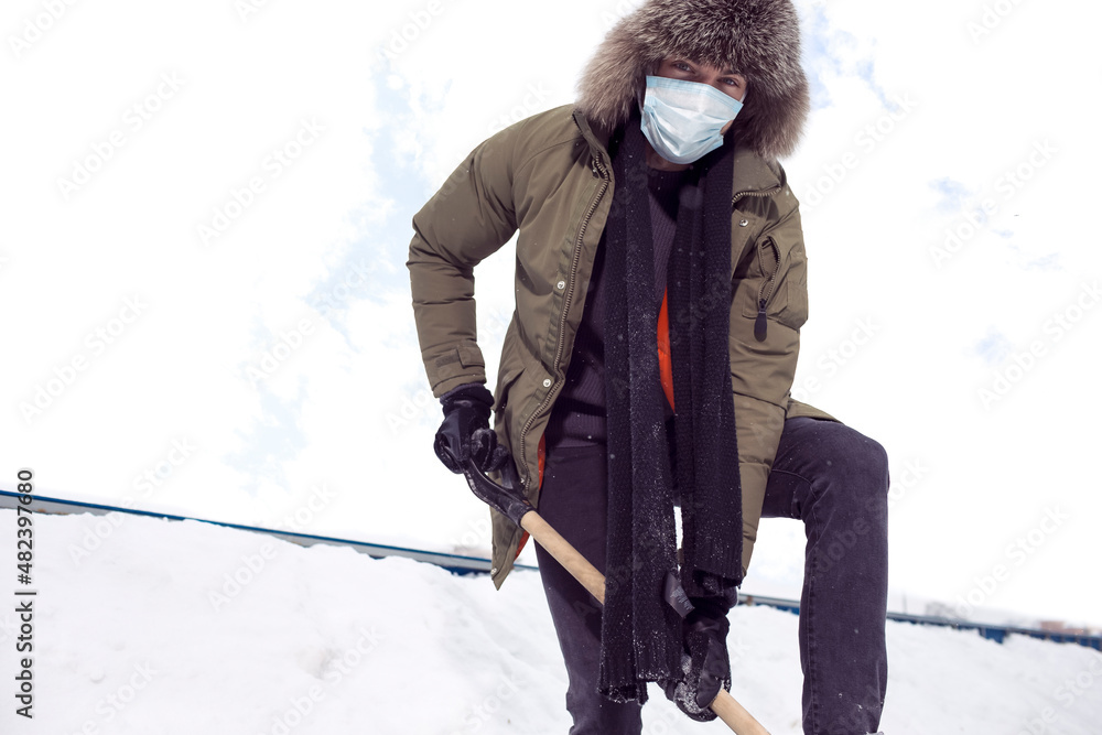 Winter Concepts. Workaholic Caucasian Handsome Man in Protective Facial Mask  Throwing Away Snow With Broken Shovel In City Against Cloudy Sky.