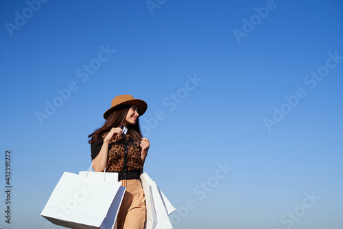 Sale or retail concept. Beautiful caucasian luxury fashionable woman in hat with shopping bags, blue sky and urban city background. Glam brunette girl in sunglasses going outdoor in sunny day