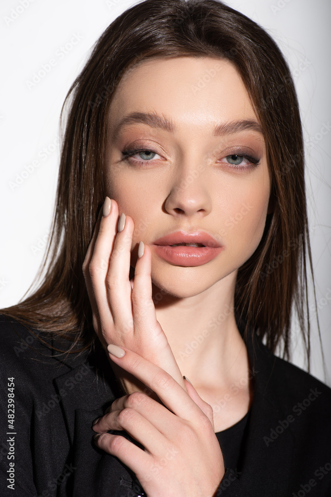 close up view of young brunette woman touching face while looking at camera isolated on grey. close up view of young brunette woman touching face while looking at camera isolated on grey.