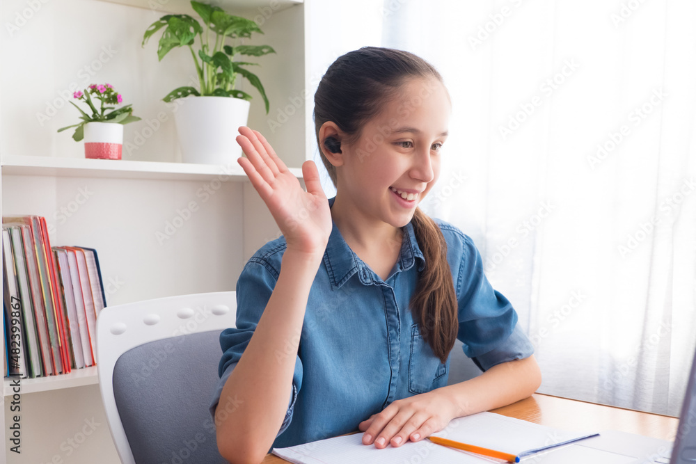 Brunette teenager in a denim shirt uses wireless headphones to study at the computer at home, studies, does homework, communicates on the Internet, waving hello. Brunette teenager in a denim shirt uses wireless headphones to study at the computer at home, studies, does homework, communicates on the Internet, waving hello.
