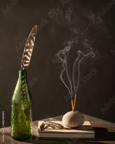 Still life with smokey incense, journal and pocket knife feather, green bottle, stones, rocks and dark background on wood table top.