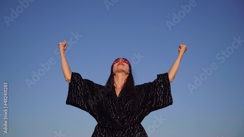 Brunette superhero girl in black dress and red face mask spreading hands with clenched fists and looking up on sky background. Female power, protest, women rights, activism concept. 4k video footage photo