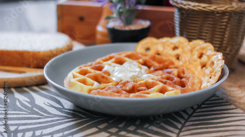 Waffles topped with honey in a gray plate with bread and seasonings placed on a wooden table.