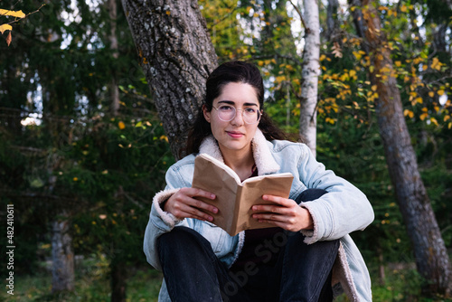A young woman with glasses reading a book sitting on a tree branch. Reading concept.