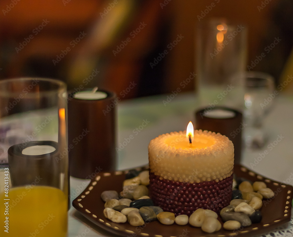 Lit decorative candle on a saucer with stones on the background of candles and glasses on the table Lit decorative candle on a saucer with stones on the background of candles and glasses on the table