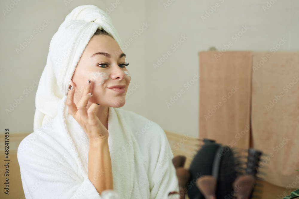 Smiling woman applying rich moisturizing face cream when standing at bathroom window after shower Smiling woman applying rich moisturizing face cream when standing at bathroom window after shower