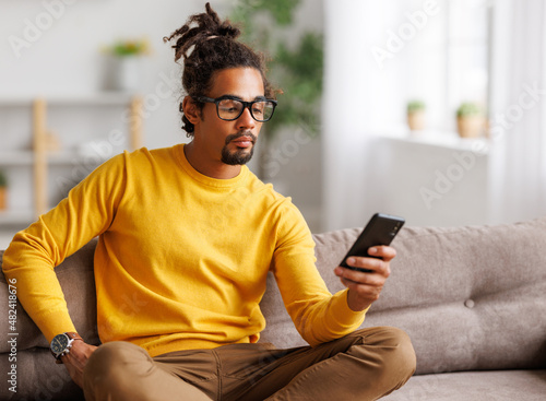 Young focused serious african american man relaxing at home with smartphone, surfing internet
