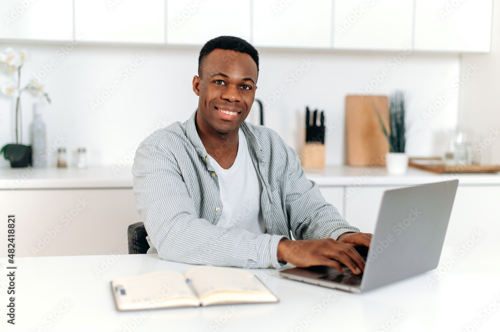 Friendly black man, freelancer, designer or student, sitting at home in the kitchen, using laptop and notebook study or surf the Internet, improving his skills, smiling friendly. E-learning concept